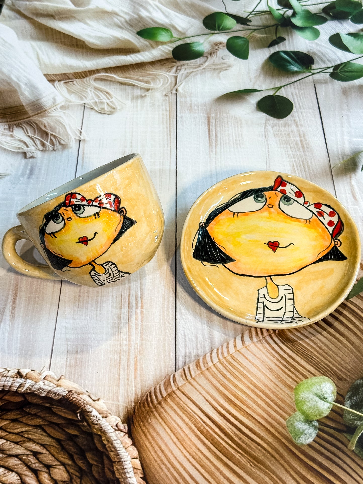 Beige ceramic mug and saucer set with hand-painted girl wearing red polka-dot headband looking up, displayed on light wooden background.
