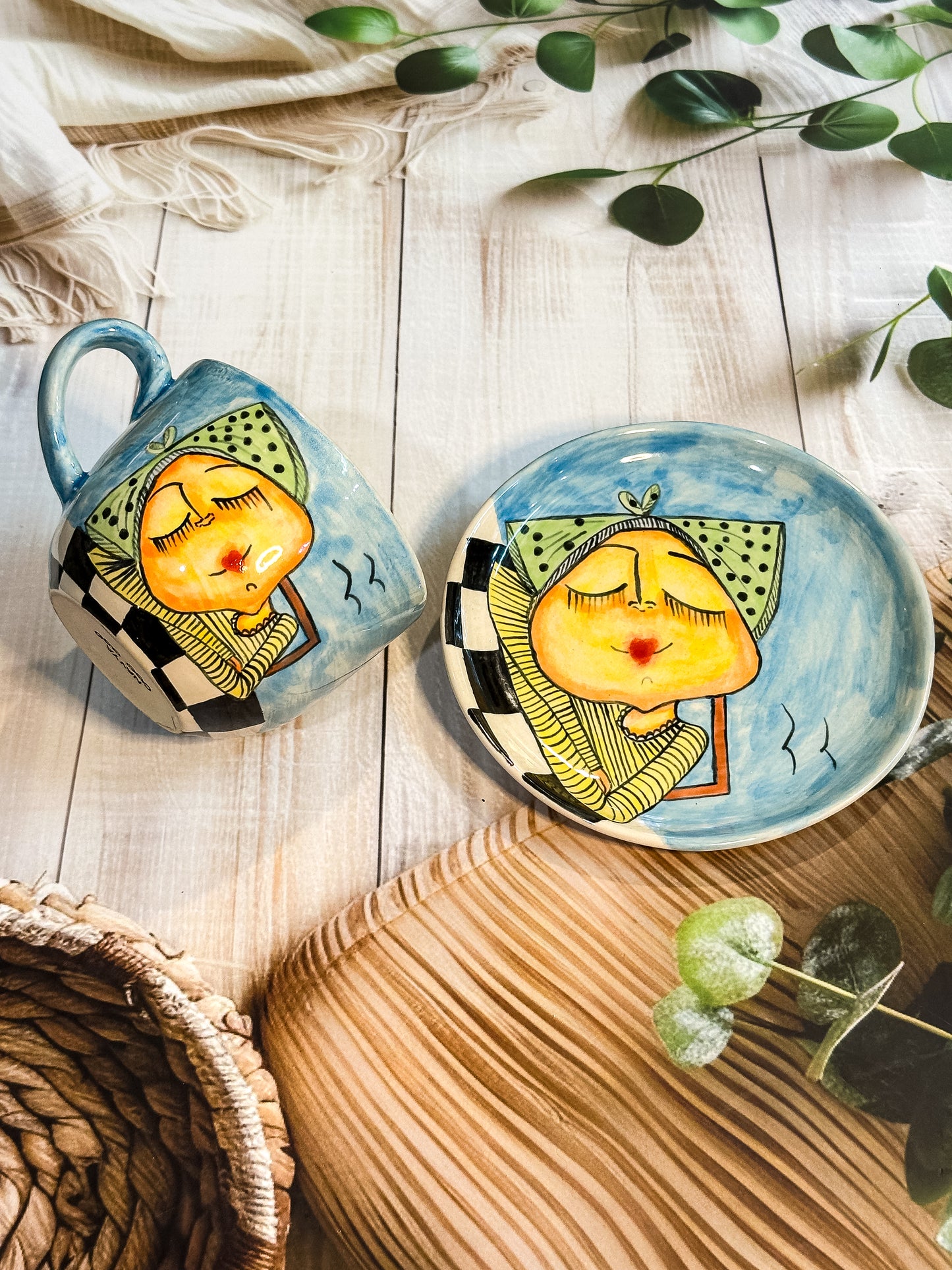 Blue hand-painted ceramic mug and saucer with whimsical girl illustration in green polka-dot headscarf blowing a kiss, styled on wooden background.