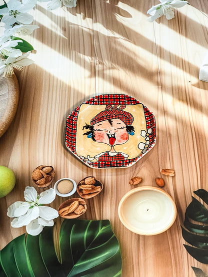 Hand-painted ceramic snack plate featuring a girl sticking her tongue out with a red checked border, styled with tropical leaves, nuts, and flowers.
