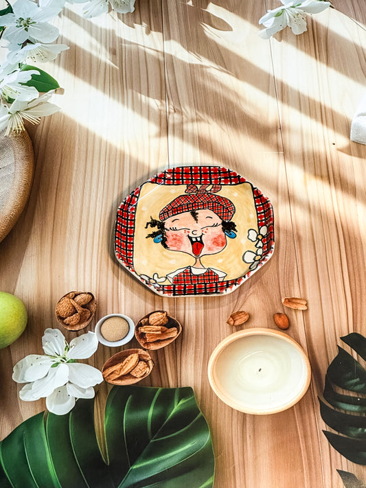 Hand-painted ceramic snack plate featuring a girl sticking her tongue out with a red checked border, styled with tropical leaves, nuts, and flowers.