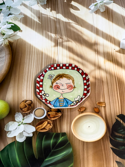 Hand-painted ceramic snack plate with a smiling girl holding daisies and a red-and-white border, styled with nuts, flowers, and a candle.
