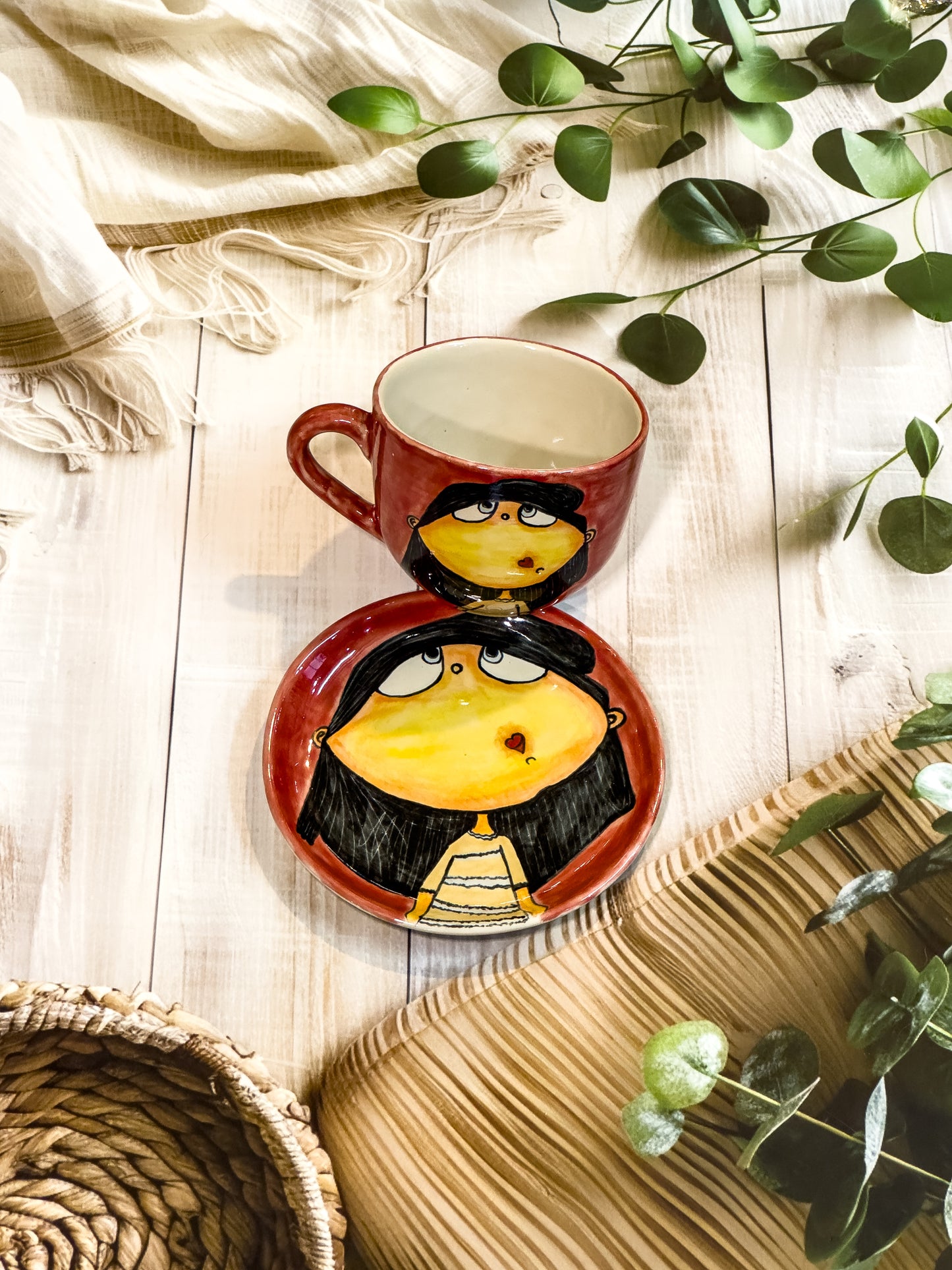 Coral red hand-painted ceramic mug and saucer featuring a wide-eyed girl with black hair and a heart-shaped mouth, styled with natural decor.