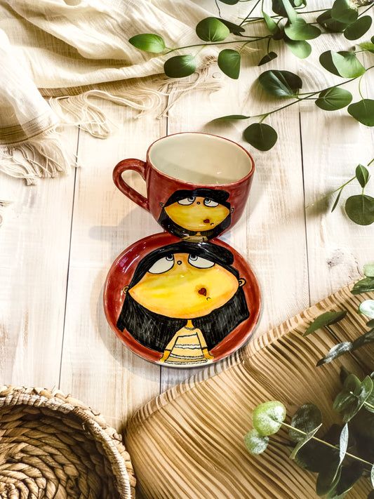Coral red hand-painted ceramic mug and saucer featuring a wide-eyed girl with black hair and a heart-shaped mouth, styled with natural decor.