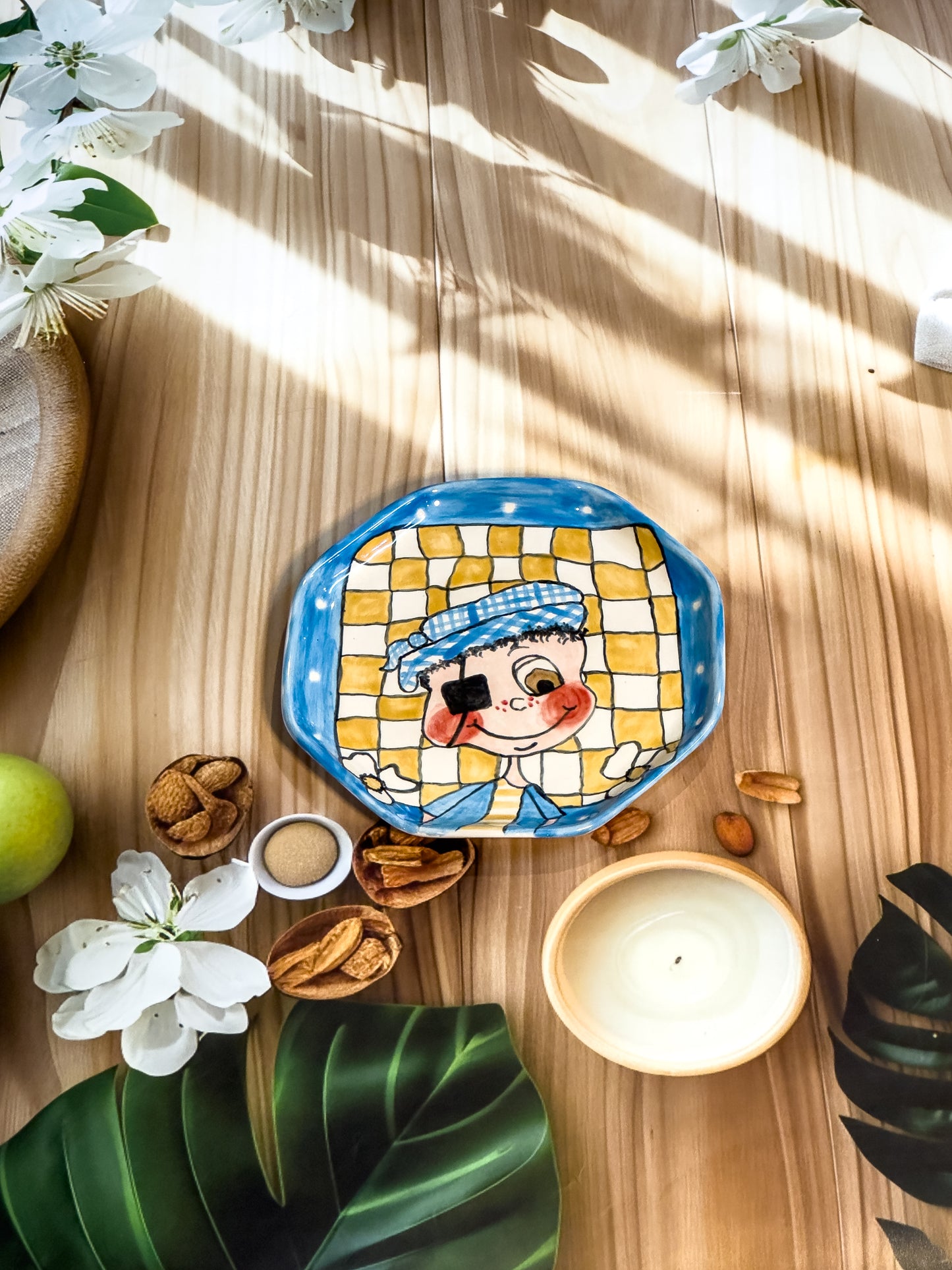 Hand-painted ceramic snack plate with a winking boy in a checked background and blue border, photographed with nuts, flowers, and a candle.