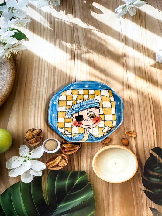 Hand-painted ceramic snack plate with a winking boy in a checked background and blue border, photographed with nuts, flowers, and a candle.