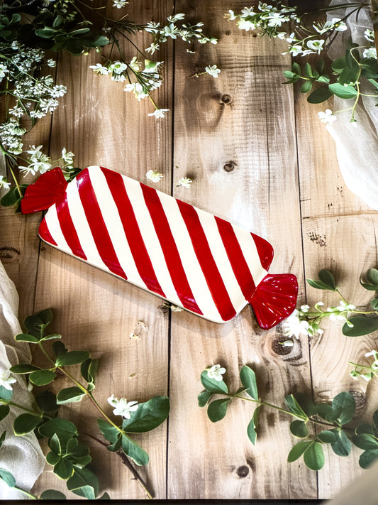 Small red and white striped handpainted ceramic toffee platter shaped like wrapped candy, artistic hostess gift on white wooden table with natural greenery

