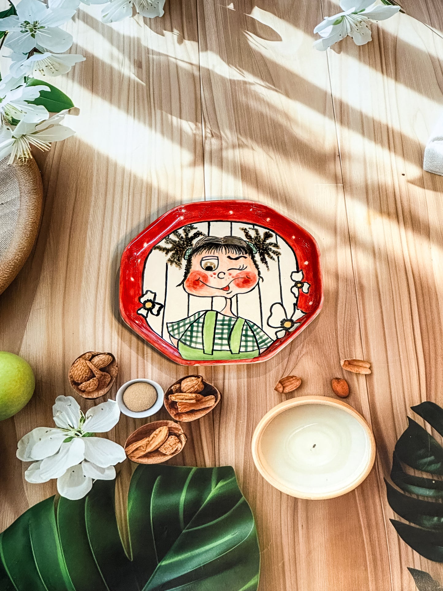Hand-painted ceramic snack plate with a winking girl with pigtails, green checks, and a red border, styled with flowers and nuts.
