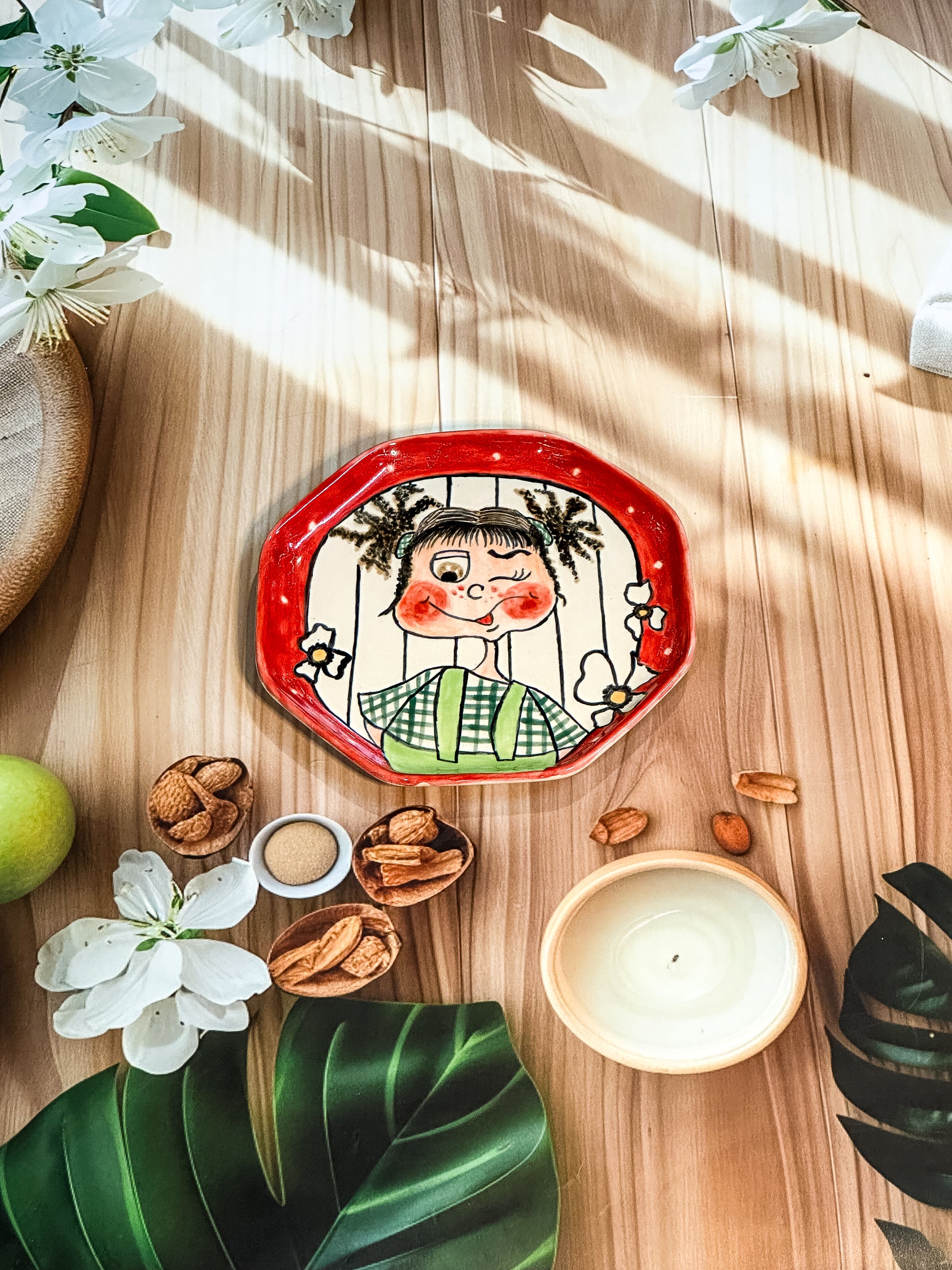 Hand-painted ceramic snack plate with a winking girl with pigtails, green checks, and a red border, styled with flowers and nuts.

